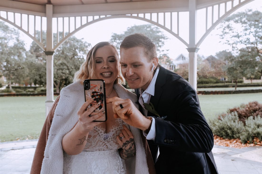 Woman in white wedding dress with veil smiling and holding a mobile phone with man in a navy jacket, smiling and pointing at the mobile phone